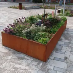 En rostfärgad blomlåda fylld med lila och vita blommor, omgiven av stenläggning.