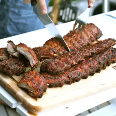 En person skär upp saftiga, grillade kamben på en skärbräda. Perfekt för en sommarkväll med grillning.