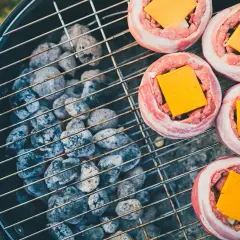Fem baconslindade köttfärsbiffar med ost på en grill för indirekt grillning. Glödande kol under gallret.