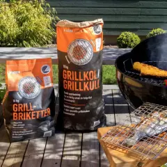 Två säckar med grillkol eller briketter står bredvid en grill med majskolvar, korvar och paprika. En fisk ligger på en grillhalster.