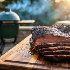 Rökig brisket skärs upp på en skärbräda. En grön kamado-grill syns i bakgrunden, perfekt för detta kamado recept.