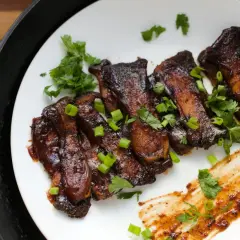 Grilla short ribs, glaserade och toppade med färsk koriander och salladslök, serverade på en vit tallrik bredvid en gjutjärnspanna.