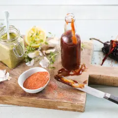 En flaska med mörk, klibbig grill glaze, en burk med grön sås, citron, vitlök och kryddor på en träskärbräda.