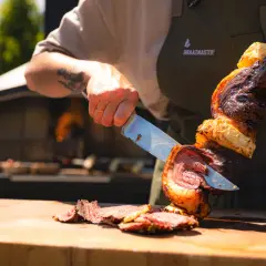 En kock skär upp en saftig bit kött från ett spett, en klassisk brasiliansk grill. Grillat kött och ananas på en träskärbräda.