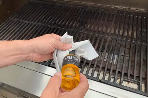 Händer oljar in grillgallret med en pappersduk och en flaska rapsolja för att maten inte ska fastna.