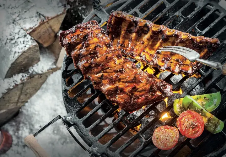 Grillade revbensspjäll med grönsaker över glödande kol. En gaffel sticker upp ur köttet. Snö och ved i bakgrunden. Perfekt för ett salt lag.