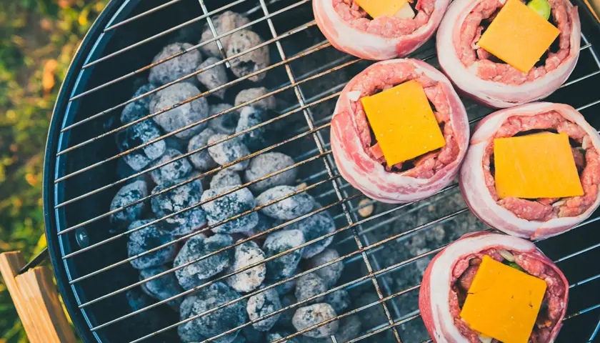 Fem baconslindade köttfärsbiffar med ost på en grill för indirekt grillning. Glödande kol under gallret.