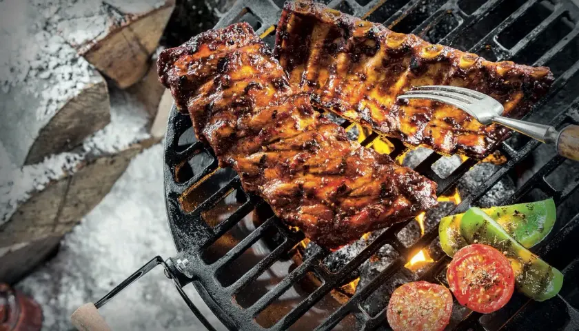 Grillade revbensspjäll med grönsaker över glödande kol. En gaffel sticker upp ur köttet. Snö och ved i bakgrunden. Perfekt för ett salt lag.