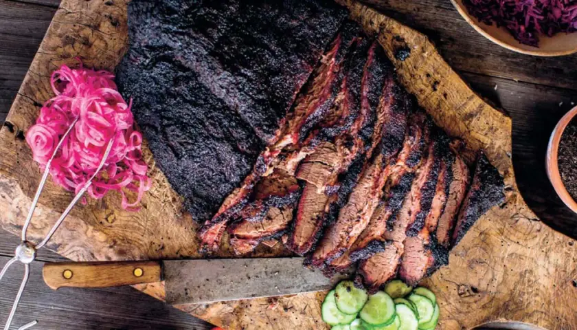 En saftig, grillad brisket skuren i skivor, serverad med picklad rödlök och gurka på en träskärbräda.