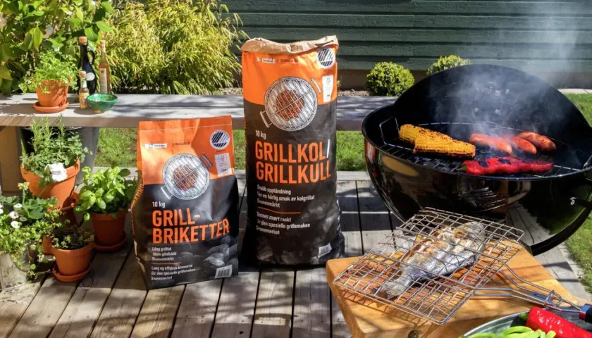 Två säckar med grillkol eller briketter står bredvid en grill med majskolvar, korvar och paprika. En fisk ligger på en grillhalster.