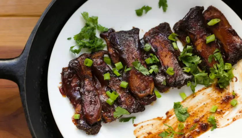 Grilla short ribs, glaserade och toppade med färsk koriander och salladslök, serverade på en vit tallrik bredvid en gjutjärnspanna.