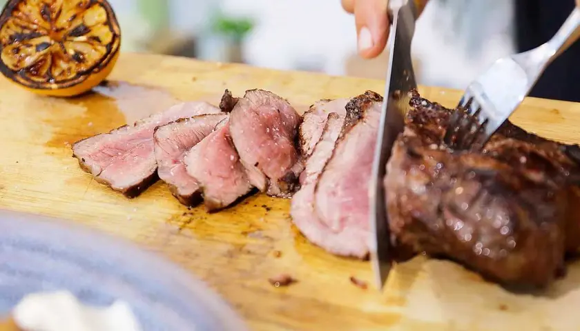 Perfekt grillad ryggbiff skärs upp på en träskärbräda. En grillad citron ligger bredvid.