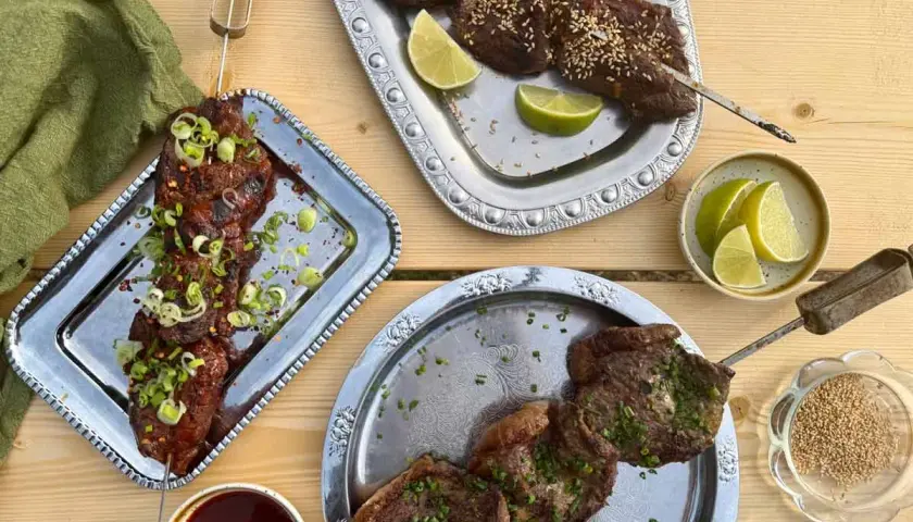 Grillad oxbringa på spett, toppad med salladslök och chiliflakes. Serveras med limeklyftor och sesamfrön.