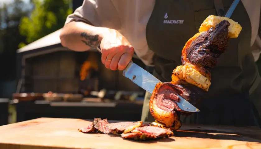 En kock skär upp en saftig bit kött från ett spett, en klassisk brasiliansk grill. Grillat kött och ananas på en träskärbräda.