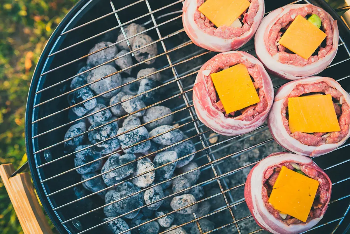 Fem baconslindade köttfärsbiffar med ost på en grill för indirekt grillning. Glödande kol under gallret.