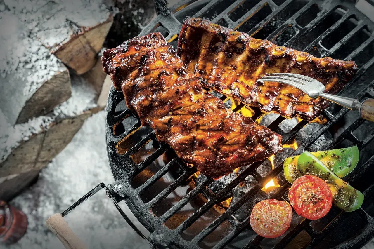 Grillade revbensspjäll med grönsaker över glödande kol. En gaffel sticker upp ur köttet. Snö och ved i bakgrunden. Perfekt för ett salt lag.