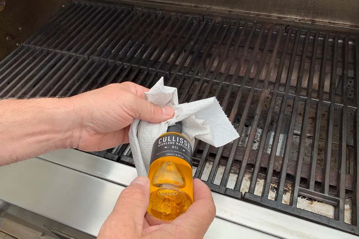 Händer oljar in grillgallret med en pappersduk och en flaska rapsolja för att maten inte ska fastna.