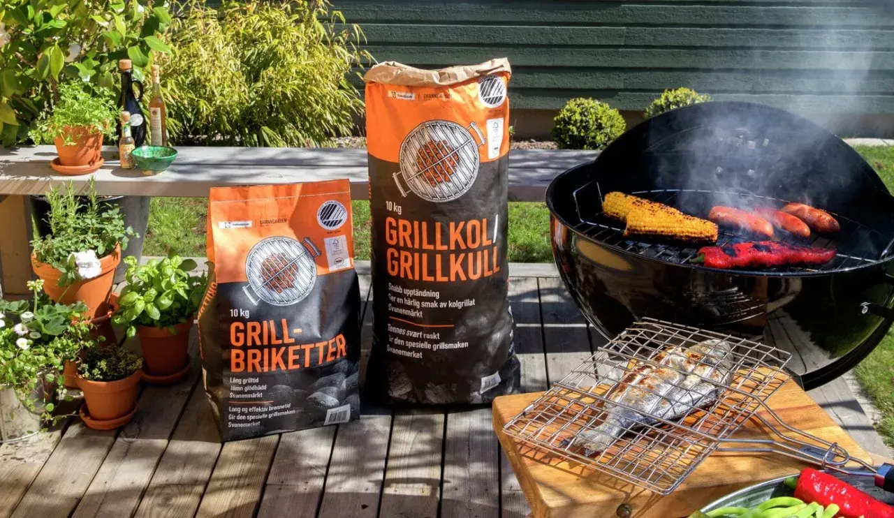 Två säckar med grillkol eller briketter står bredvid en grill med majskolvar, korvar och paprika. En fisk ligger på en grillhalster.