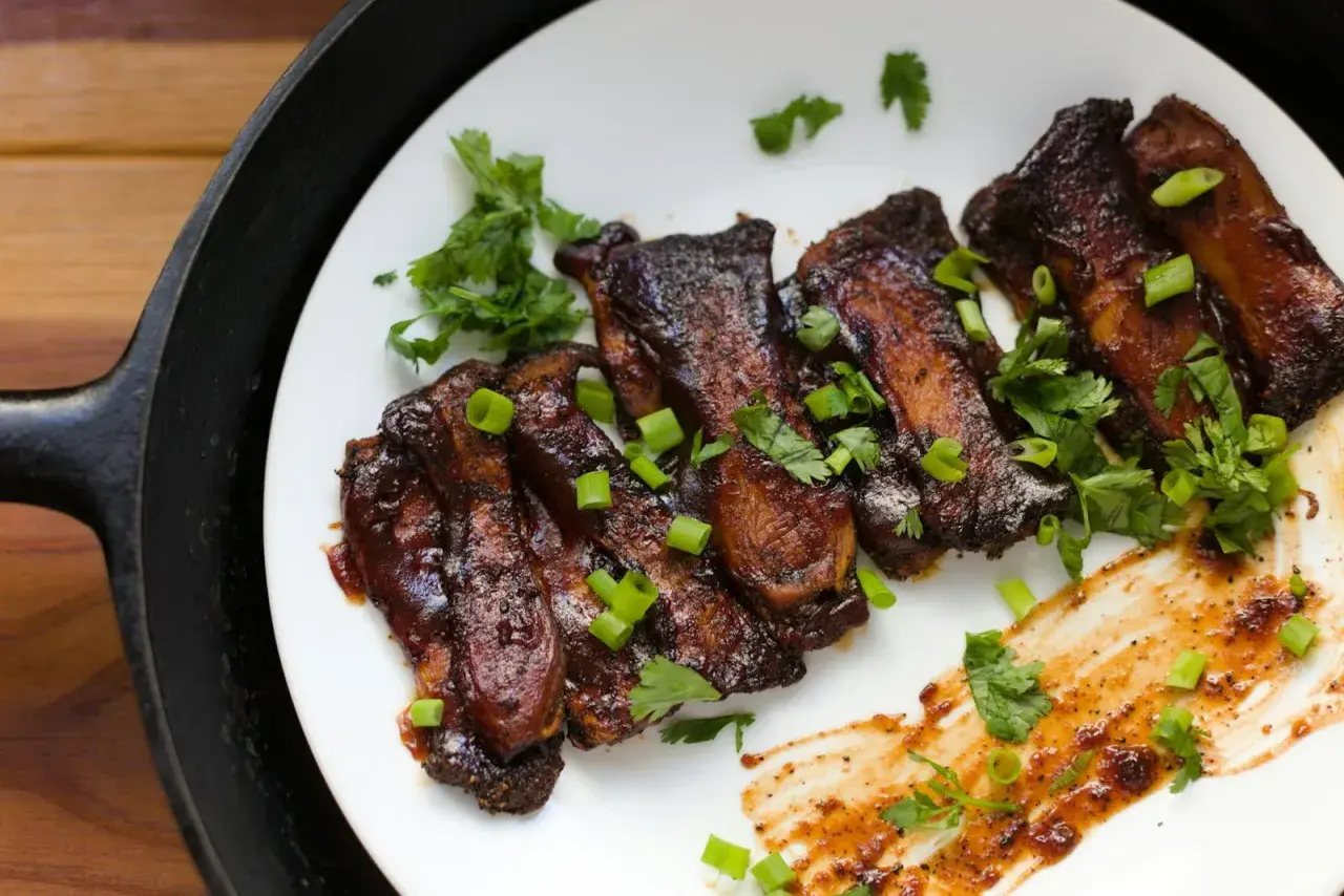 Grilla short ribs, glaserade och toppade med färsk koriander och salladslök, serverade på en vit tallrik bredvid en gjutjärnspanna.