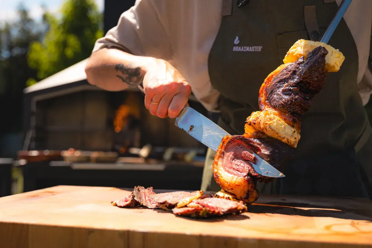 En kock skär upp en saftig bit kött från ett spett, en klassisk brasiliansk grill. Grillat kött och ananas på en träskärbräda.