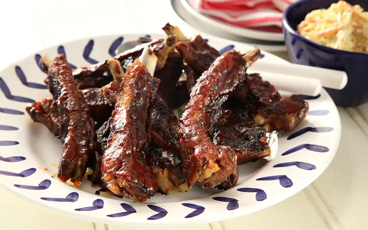 En tallrik med glaserade, saftiga marinad spareribs. Bredvid syns en sk&aring;l med kr&auml;mig coleslaw.