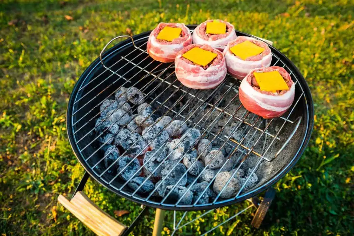 Grilla med kolgrill: fem baconsnurrade hamburgare med ost p&aring; en gl&ouml;dande kolgrill i gr&auml;set.