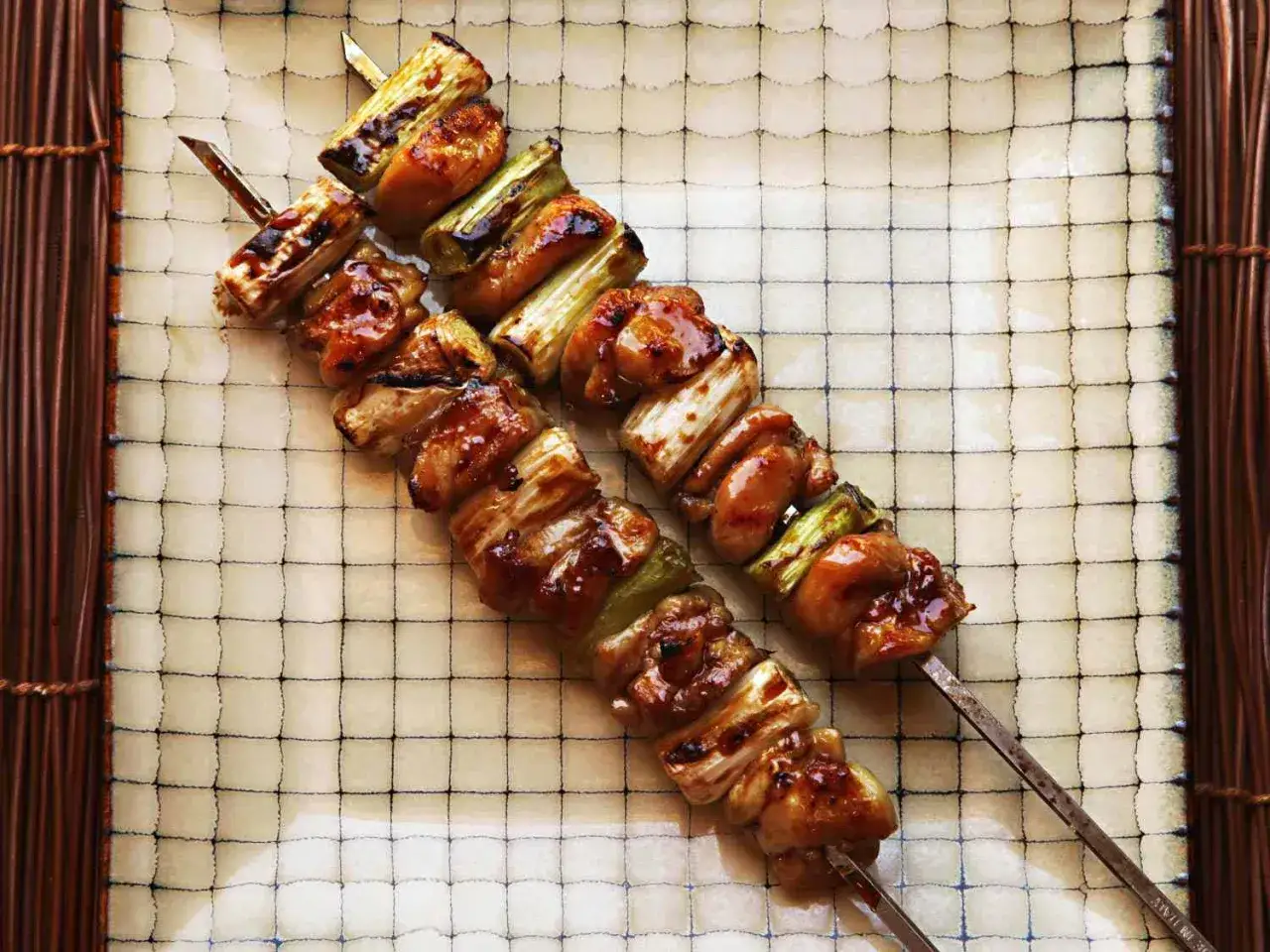 Två saftiga yakitori kycklingspett med glaserad kyckling och gröna lökar, serverade på en rutig tallrik.