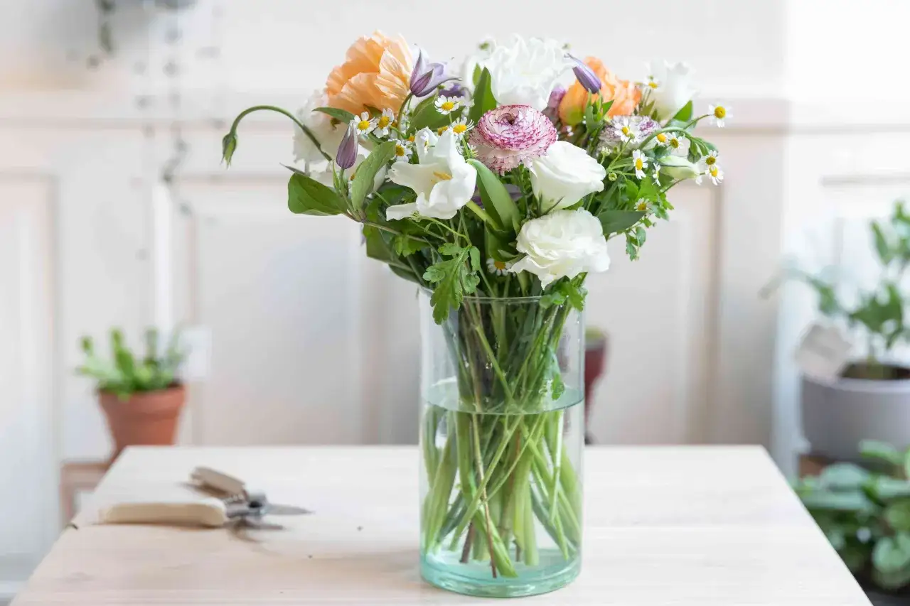 En bukett med vanliga snittblommor i en glasvas, med en blomstersax p&aring; bordet.