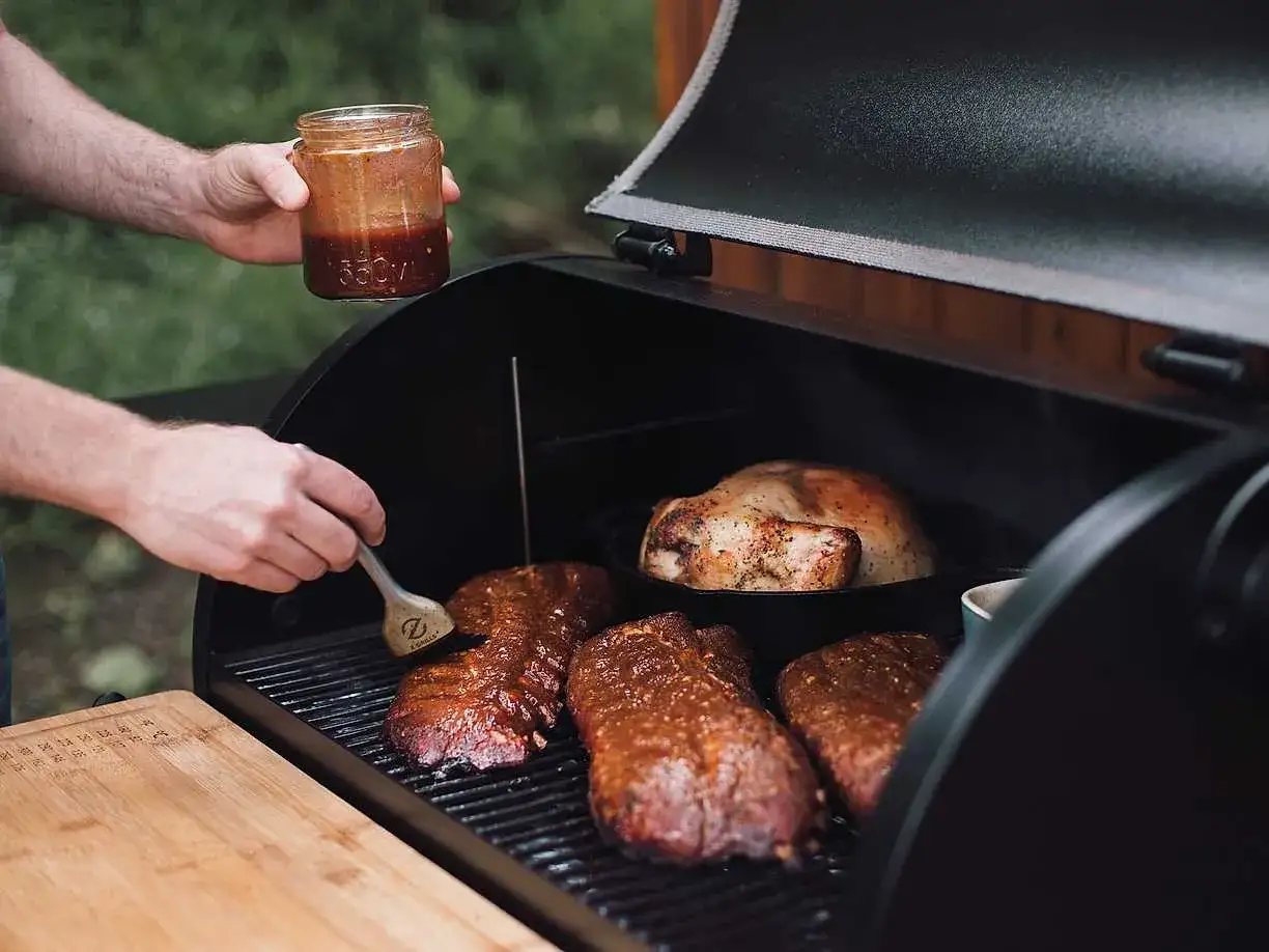 En person penslar revbensspj&auml;ll med en h&auml;rlig revbens glaze p&aring; grillen. En hel kyckling steks bredvid.