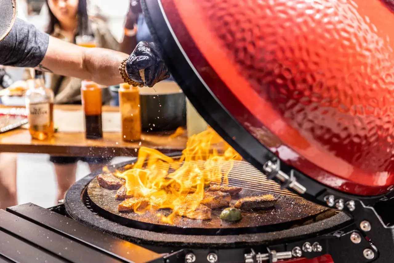 K&ouml;tt steks p&aring; en kamado grill med flammor. En hand med handske droppar en s&aring;s &ouml;ver k&ouml;ttet. Ett perfekt kamado recept!