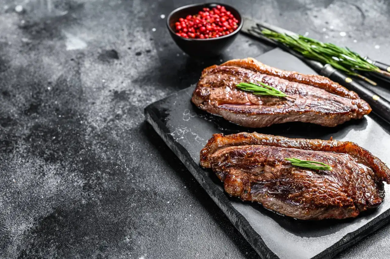 Tv&aring; perfekt grillade picanha stekar med rosmarin p&aring; en svart stenplatta. En sk&aring;l med r&ouml;da pepparkorn och en gaffel syns i bakgrunden.